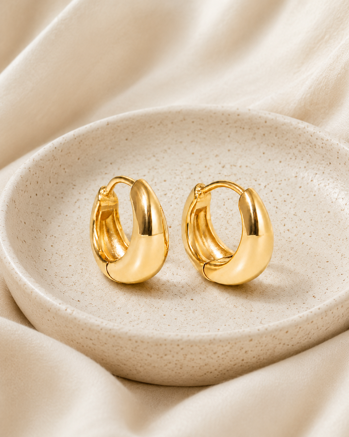 Bold gold hoop earrings in a ceramic dish on soft ivory fabric.